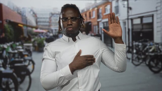 Man wearing white shirt and glasses with hand on chest and right hand raised in street with bicycles and brick buildings; pledge solemn.