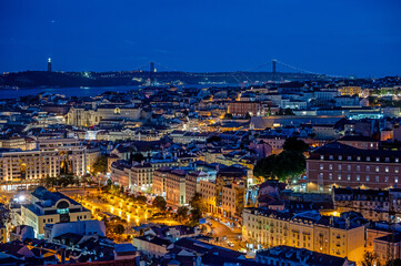 Naklejka premium Illuminated buildings of Lisbon, Portugal in deep afternoon twilight with Tagus River and bridge in background from Miradouro da Senhora do Monte.
