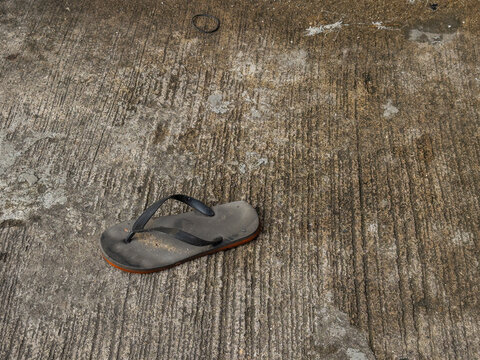 A single, worn black flip-flop with an orange sole lies discarded on a rough, textured concrete surface under natural light. This everyday object evokes themes of simplicity, loss, or a temporary