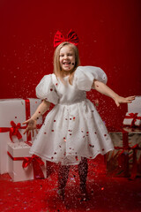 little girl in an elegant white dress on a red background in a studio with gift boxes catches confetti