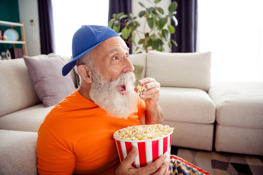Funny grandpa enjoys popcorn at home wearing an orange shirt and blue cap while relaxing on a sofa indoors
