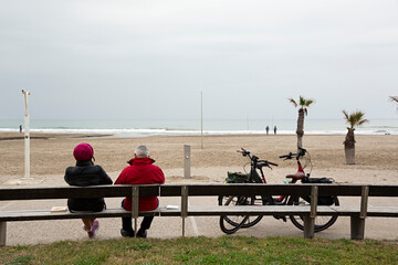 Paisaje de playa con pareja sentada en un banco y sus bicicletas.