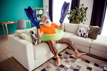Funny grandpa frolics indoors with inflatable ring and blue fins in cozy homey living room