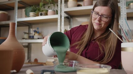 Woman pouring green paint into a container in a ceramics workshop. Preparing glaze for painting handmade clay pots, artisan working in a pottery studio