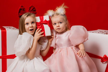 two little girls in elegant white dresses on a red background in a studio with gift boxes catching confetti for the New Year