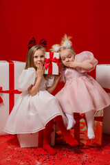 two little girls in elegant white dresses on a red background in a studio with gift boxes catching confetti for the New Year