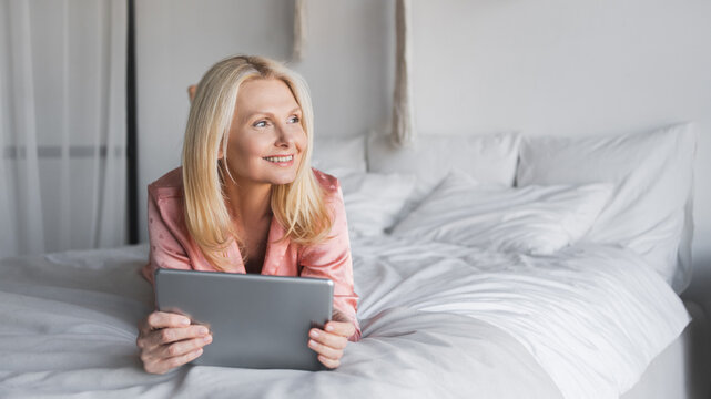 Middle age woman lying on stomach on bed looking on copy space in bedroom, holding tablet, smiling. Remote working, technology, lifestyle, minimalist, interior, natural light, relaxation