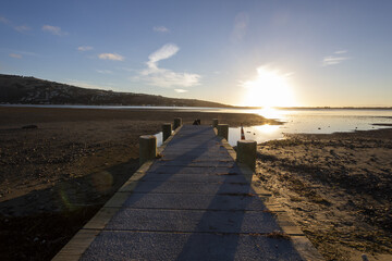 View of a weathered wooden pier stretching out into the calm waters under a bright sun, casting long shadows on the shore, Christchurch, Canterbury Region, New Zealand.