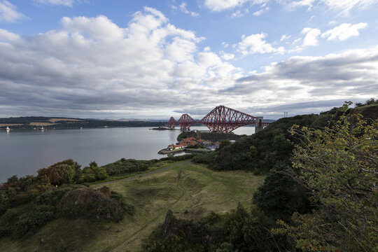 View of the iconic red Forth Bridge dramatically spans the Firth of Forth under a sky filled with swirling clouds, set against the lush green coastline, Edinburgh, Scotland, United Kingdom.