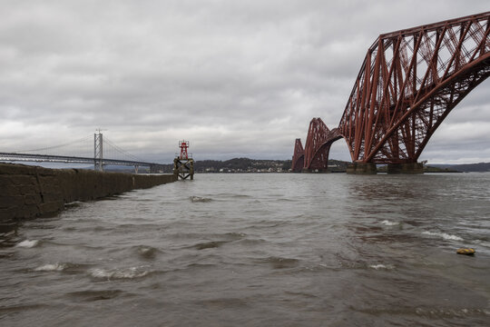 View of the iconic red Forth Bridge dramatically spans the Firth of Forth under a moody sky, waves gently lapping against the shore, Edinburgh, Scotland, United Kingdom.