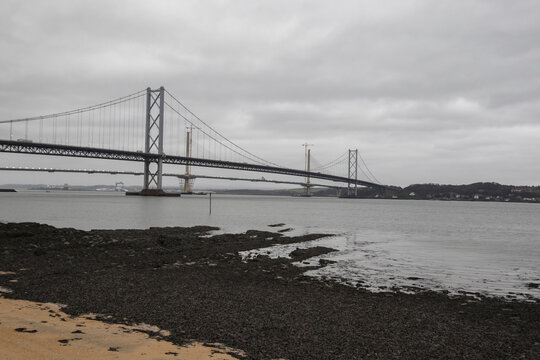 View of the majestic Forth Road Bridge stretches across the calm water under a brooding sky, its steel cables a stark contrast to the dark, rocky shore, Edinburgh, Scotland, United Kingdom.