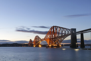 View of the iconic Forth Bridge ablaze with warm light reflecting on the tranquil waters under a dusky sky, Edinburgh, Scotland, United Kingdom.