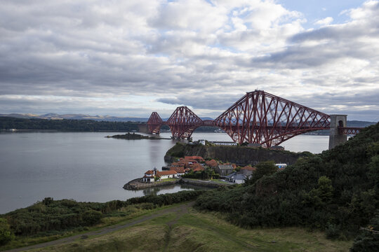 View of the iconic red Forth Bridge stretches across the calm waters under a sky of textured clouds, connecting distant shores, Edinburgh, Scotland, United Kingdom.