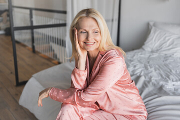 Smiling blond haired mature woman middle age female wearing pink sleepwear sitting on bed, in cozy room after good dreams, morning sunlight, wellness, copy space. Hotel apartment concept