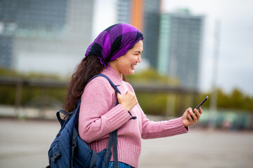 Smiling Asian woman with backpack using her phone in urban environment