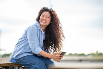 Smiling young Asian woman with curly hair holding smartphone while sitting outdoors