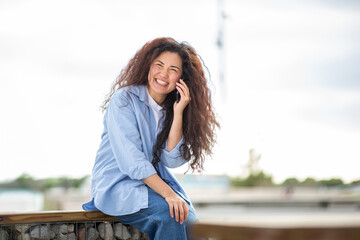 Joyful young Asian woman talking on phone while sitting in urban outdoor setting