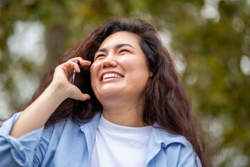 Happy Asian woman with curly hair talking on smartphone in park