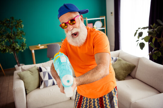Funny grandpa with blue cap plays with water gun indoors in bright living room wearing orange shirt and striped shorts smiling and having a fun home moment