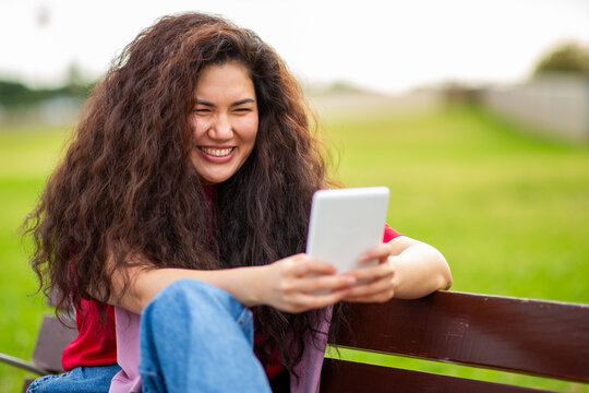 Smiling Asian woman reading an ebook while sitting on park bench in casual outfit - Powered by Adobe