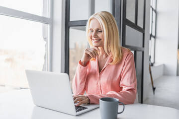 Happy mature business woman sitting at home office table drinking coffee looking at laptop. Smiling middle aged female freelancer working remotely on computer checking email in modern house.