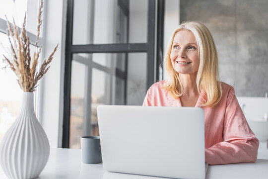 Happy aged woman working at laptop drinking morning tea coffee, smiling mature female using computer browsing or surfing internet, reading news online, excited lady texting message at pc at home - Powered by Adobe