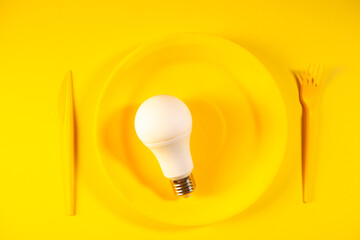 Light bulb on yellow plate with fork and knife on yellow background, top view