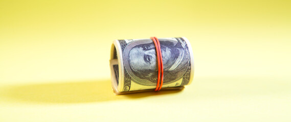 Roll of one hundred dollar bills tied with a rubber band on a yellow background.