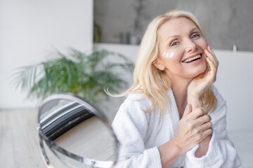 Happy, reflection and mature person with cosmetics application for glowing face in home. Beauty,...