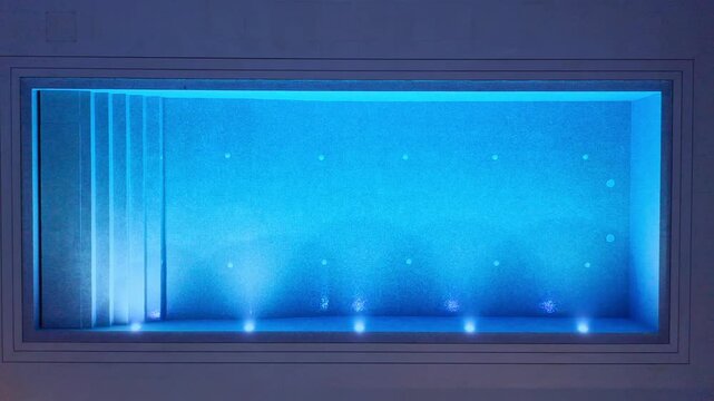 Descending drone view of modern rectangular swimming pool at night illuminated with blue LED lights showing steps and underwater lighting pattern Concept of luxury relaxation and nighttime ambiance