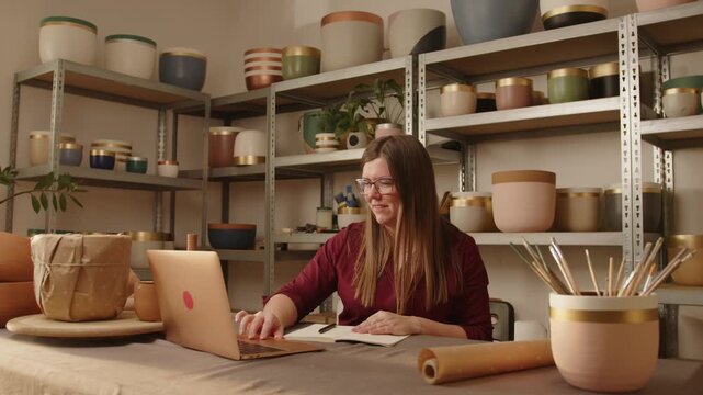 Woman working on a laptop and writing notes in a notebook inside a ceramics workshop. Small business owner managing orders and planning production among handmade pottery