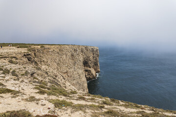 View of rugged cliffs meet the vast, deep blue sea under a hazy sky, a serene yet dramatic coastal scene unfolds, , Faro, Algarve, Portugal.