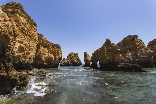 View of golden cliffs meet the turquoise sea, waves crash against the rocky shore under a clear blue sky, a mesmerizing coastal scene, Faro, Algarve, Portugal. - Powered by Adobe