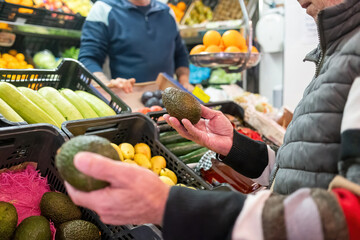 An older man carefully selects avocados at a fruit stand, surrounded by colorful produce in a busy market environment.