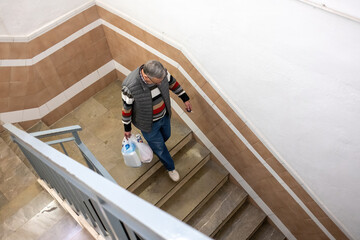 An older man descends a stairwell carrying grocery bags, captured in a natural everyday moment inside a residential building.