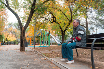 An older man sits on a park bench near a playground, enjoying a quiet break amid colorful autumn trees and soft natural light.