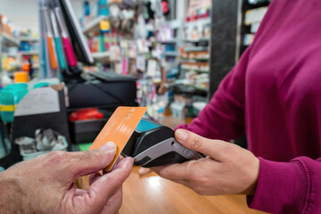 Senior man makes a contactless card payment while a shop worker holds the terminal in a small retail store filled with everyday products.