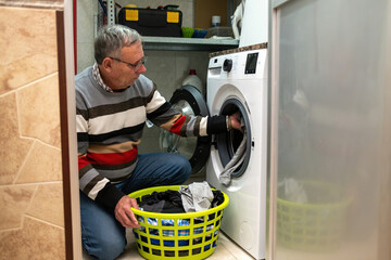 Un hombre mayor se arrodilla en el cuarto de lavado metiendo ropa en la lavadora mientras sostiene un cesto lleno, realizando una tarea dom stica cotidiana.