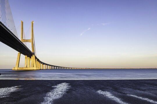 View of the Vasco da Gama Bridge stretches across the water, its yellow pillars contrasting against the soft blue sky, Lisbon, Vasco da Gama, Portugal.