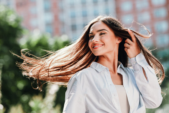 Playful sunny urban style woman posing in white shirt with flowing hair smiling in city park scene for fashion lifestyle stock photo