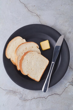 Three (3) slices of Jamaican hard dough bread on a black plate with a knife and a cube of butter