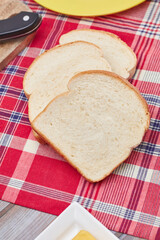 Three slices of Jamaican hard dough bread on a Jamaican bandana with a yellow plate, cutting board, knife and a stick of butter