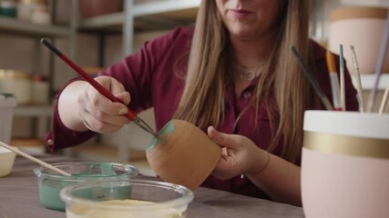 Woman painting a handmade clay pot in a ceramics workshop. Artisan applying green paint with a brush, crafting and decorating pottery in an art studio