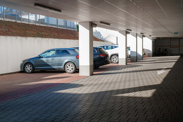 Cars parked in a covered parking area with shadows creating a pattern on the pavement, offering...