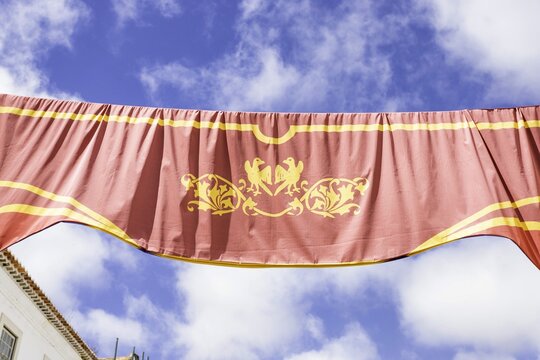 View of a red and yellow banner adorned with ornate golden emblems arches across a bright blue sky dotted with fluffy white clouds, Obidos, Leiria District, Portugal.