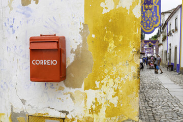 View of a vibrant red 'CORREIO' postbox affixed to a weathered wall of peeling yellow paint, contrasting with the distant street scene, Obidos, Leiria District, Portugal.