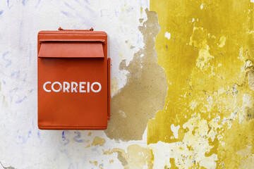 View of a vibrant red 'CORREIO' mailbox clinging to a weathered wall where layers of yellow and white paint peel away, Obidos, Leiria District, Portugal.