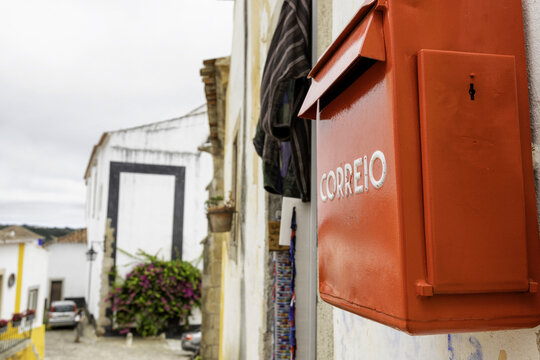 View of a vibrant red 'CORREIO' mailbox clinging to a textured white wall, contrasting with the distant, blurred medieval buildings, Obidos, Leiria District, Portugal.