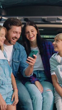 Cheerful lovely parents with cute teen kids which using mobile phone sitting in the car's trunk. Mother Father and daughter son surfing internet online on cellphone. Family road trip with trailer car