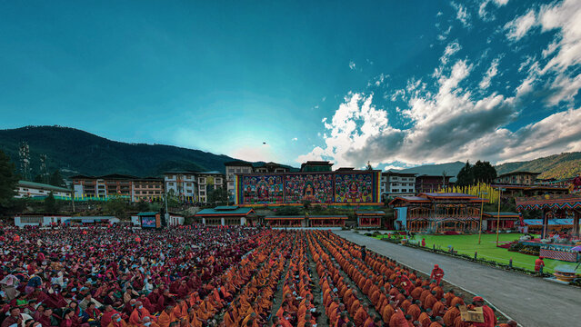 Aerial view of monks gathered in vibrant orange robes before the ornate Tashichho Dzong under a vast sky, Thimphu, Thimphu, Bhutan.